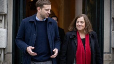 Ein Mann im blauen Mantel gestikuliert neben einer Frau mit rotem Schal vor einem Gebäude.