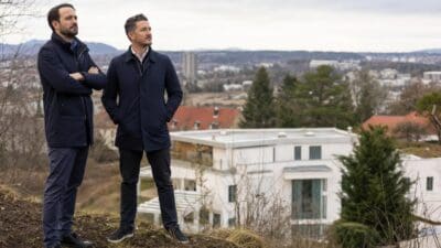 Zwei Männer in dunklen Jacken stehen auf einem Hügel vor einem modernen weißen Gebäude mit Blick auf die Stadt.