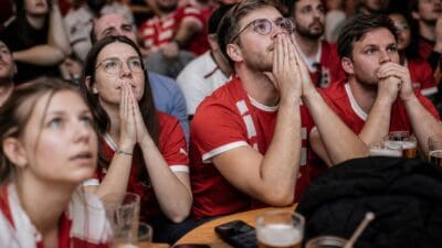 Zuschauer in roten Trikots verfolgen gespannt das Fußballspiel an einem Tisch mit Biergläsern