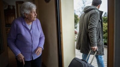 Ältere Frau in lila Strickjacke steht in Tür und schaut einem Mann mit Koffer nach