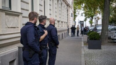 Drei Polizisten in Uniform stehen auf einem Gehweg vor einem Gebäude und beobachten eine Gruppe Menschen.