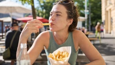 Eine Frau isst Pommes frites aus einer Papiertüte an einem Tisch im Freien.