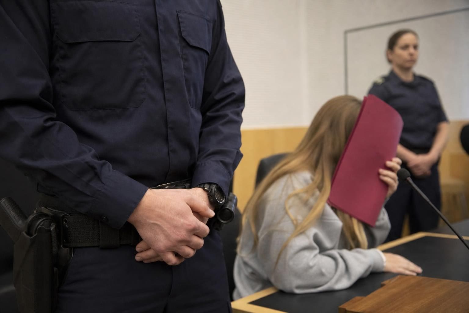Polizist im Gerichtssaal neben einer jungen Frau, die ihr Gesicht mit einer roten Mappe verdeckt