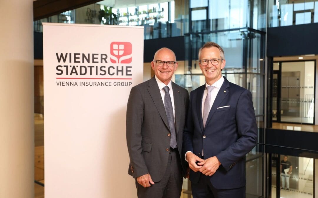 Zwei Männer in Anzügen stehen vor einem Wiener Städtische Banner in einem Bürogebäude