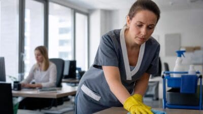Reinigungskraft in grauer Uniform und gelben Handschuhen wischt einen Tisch in einem Büro ab