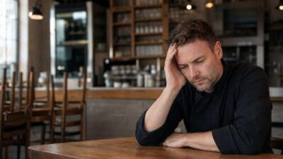 Ein Mann in Kochjacke sitzt mit der Hand am Kopf an einem Tisch in einem Restaurant