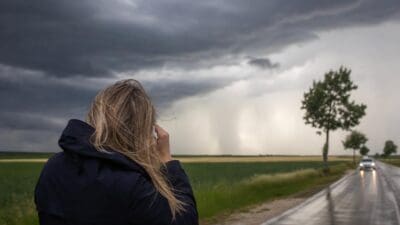 Frau mit blonden Haaren in blauer Jacke blickt auf dunkle Gewitterwolken über nasser Landstraße