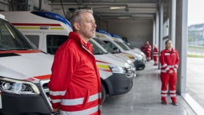 Mann in roter Einsatzjacke steht vor mehreren Rettungswagen in einer Fahrzeughalle