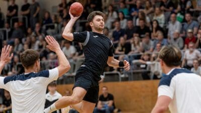 Handballspieler in schwarzem Trikot springt mit Ball in Sporthalle, Gegner in Weiß und Zuschauer im Hintergrund