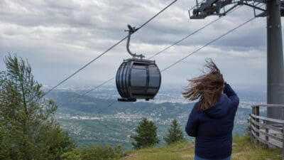 Frau mit wehendem Haar steht vor einer Seilbahngondel auf einem Berg bei Graz