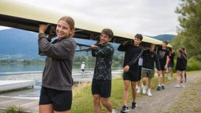 Jugendliche tragen gemeinsam ein langes Ruderboot auf den Schultern am Seeufer