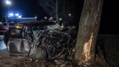 Schwer beschädigtes Auto nach Unfall gegen Baum in der Nacht mit Rettungskräften im Hintergrund