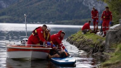 Rettungskräfte in roten Anzügen bergen verletzte Person auf Paddleboard aus dem Wasser