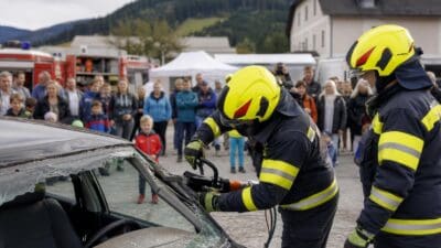 Zwei Feuerwehrmänner in gelben Helmen bearbeiten die Frontscheibe eines beschädigten Autos bei einer Übung.