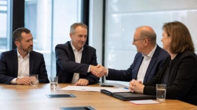 Vier Geschäftsleute in Anzügen sitzen an einem Konferenztisch, zwei Männer schütteln sich die Hände.