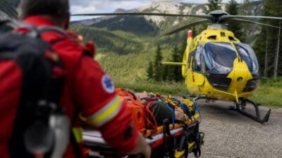 Rettungskraft in roter Uniform neben Trage mit Patienten vor gelbem Hubschrauber in den Bergen
