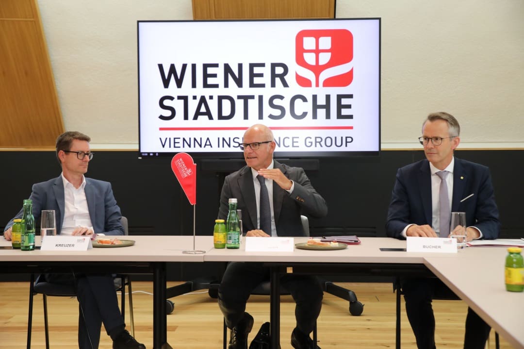 Drei Männer in Anzügen sitzen an einem Tisch vor einem Bildschirm der Wiener Städtischen
