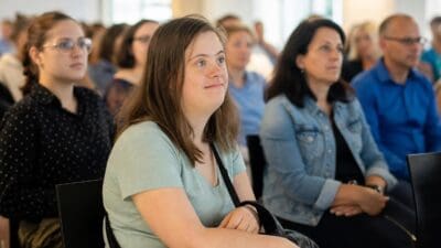 Junge Frau mit Down-Syndrom sitzt aufmerksam in einem Konferenzraum während einer Tagung.
