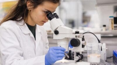 Wissenschaftlerin in weißem Kittel blickt durch Mikroskop und hält Pinzette in der Hand