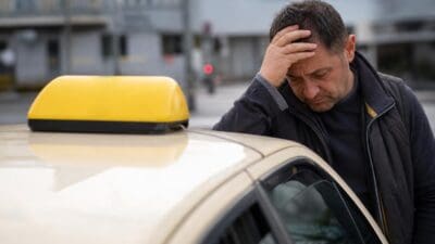 Verzweifelter Taxifahrer stützt mit der Hand den Kopf ab und lehnt an einem Taxi