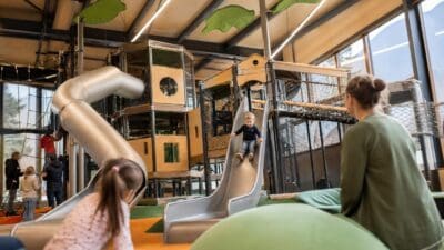 Kinder spielen auf Rutschen und Klettergerüsten in einem hellen Indoor-Spielplatz mit großen Fenstern