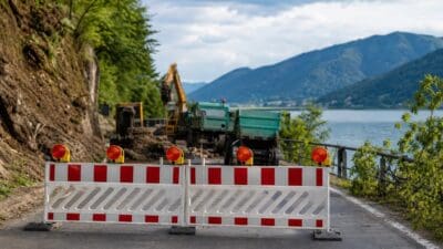 Rot-weiß gestreifte Absperrschranken mit Warnleuchten blockieren eine Straße am Seeufer