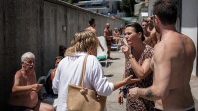 Frau im geblümten Kleid gestikuliert vor shirtlosen Männern an Betonmauer im Strandbad
