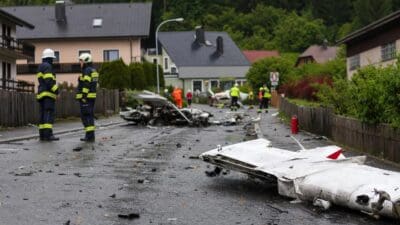 Trümmer eines Ultraleichtflugzeugs liegen auf einer nassen Straße, Rettungskräfte stehen in der Nähe von Wohnhäusern.