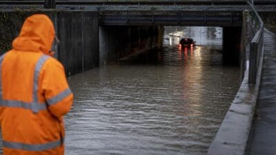 Eine Person in orangefarbener Warnjacke beobachtet ein Auto, das durch das überflutete Wasser einer Unterführung fährt.