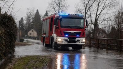 Rotes Feuerwehrfahrzeug mit Blaulicht fährt durch überflutete Straße bei Ludmannsdorf