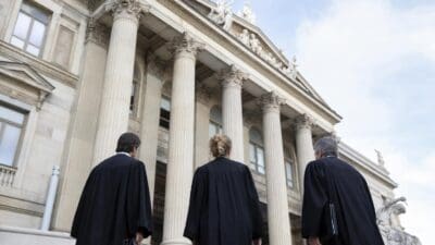 Drei Personen in schwarzen Richterroben stehen vor einem klassizistischen Gerichtsgebäude mit Säulen