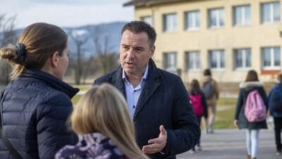 Ein Mann im dunklen Mantel spricht mit einer Frau und einem Kind vor einem Schulgebäude