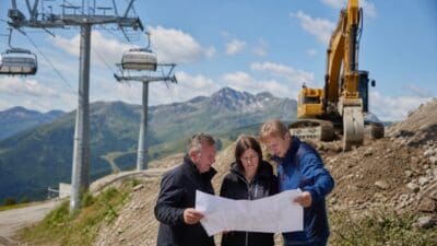 Drei Personen betrachten Baupläne vor einem Bagger und einer Seilbahn am Berg.