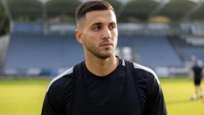 Junger Mann mit kurzen dunklen Haaren und Bart in schwarzem Sporttrikot auf grünem Rasen vor leerer Tribüne