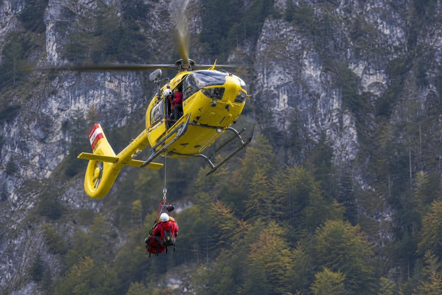 Gelber Rettungshubschrauber mit angehängter Trage über bewaldetem Berggelände