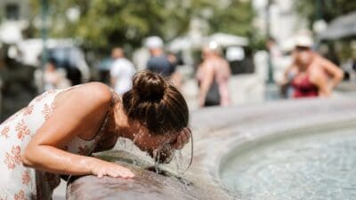 Eine Frau mit hochgestecktem Haar bespritzt sich das Gesicht mit Wasser aus einem Brunnen.