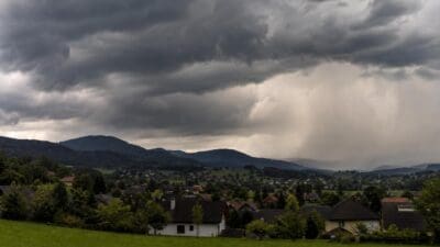 Dorf mit Häusern und grünen Wiesen vor Hügeln unter schweren dunklen Gewitterwolken