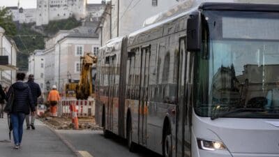 Weißer Gelenkbus fährt an Baustelle mit Bagger und Fußgängern vorbei