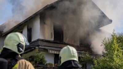 Zwei Feuerwehrleute mit Helmen betrachten das verrauchte Wohnhaus von hinten