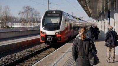 Roter und weißer Zug fährt auf einen Bahnsteig mit wartenden Personen zu