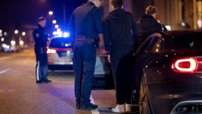 Polizisten kontrollieren zwei junge Männer neben einem Auto bei Nacht auf der Straße