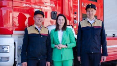 Zwei Männer in Feuerwehruniform mit einer Frau in grünem Anzug vor rotem Feuerwehrfahrzeug