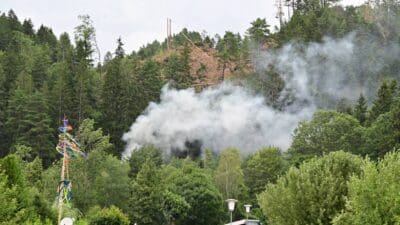 Rauch steigt von einem bewaldeten Hang auf, im Vordergrund steht ein geschmückter Maibaum.