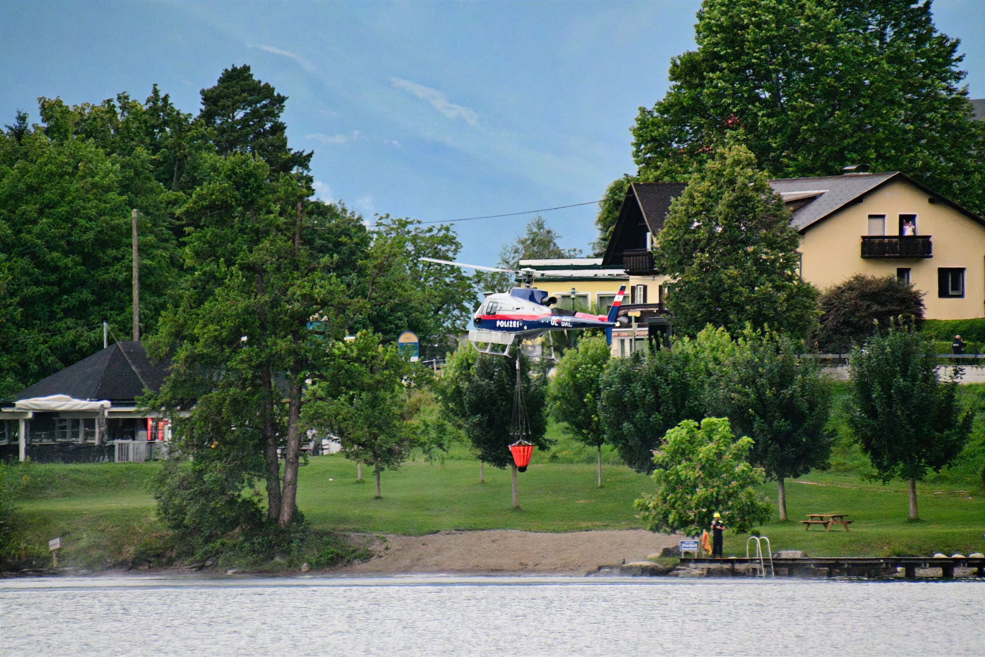 Polizeihubschrauber mit rotem Löschbehälter fliegt über Ufer am Millstätter See