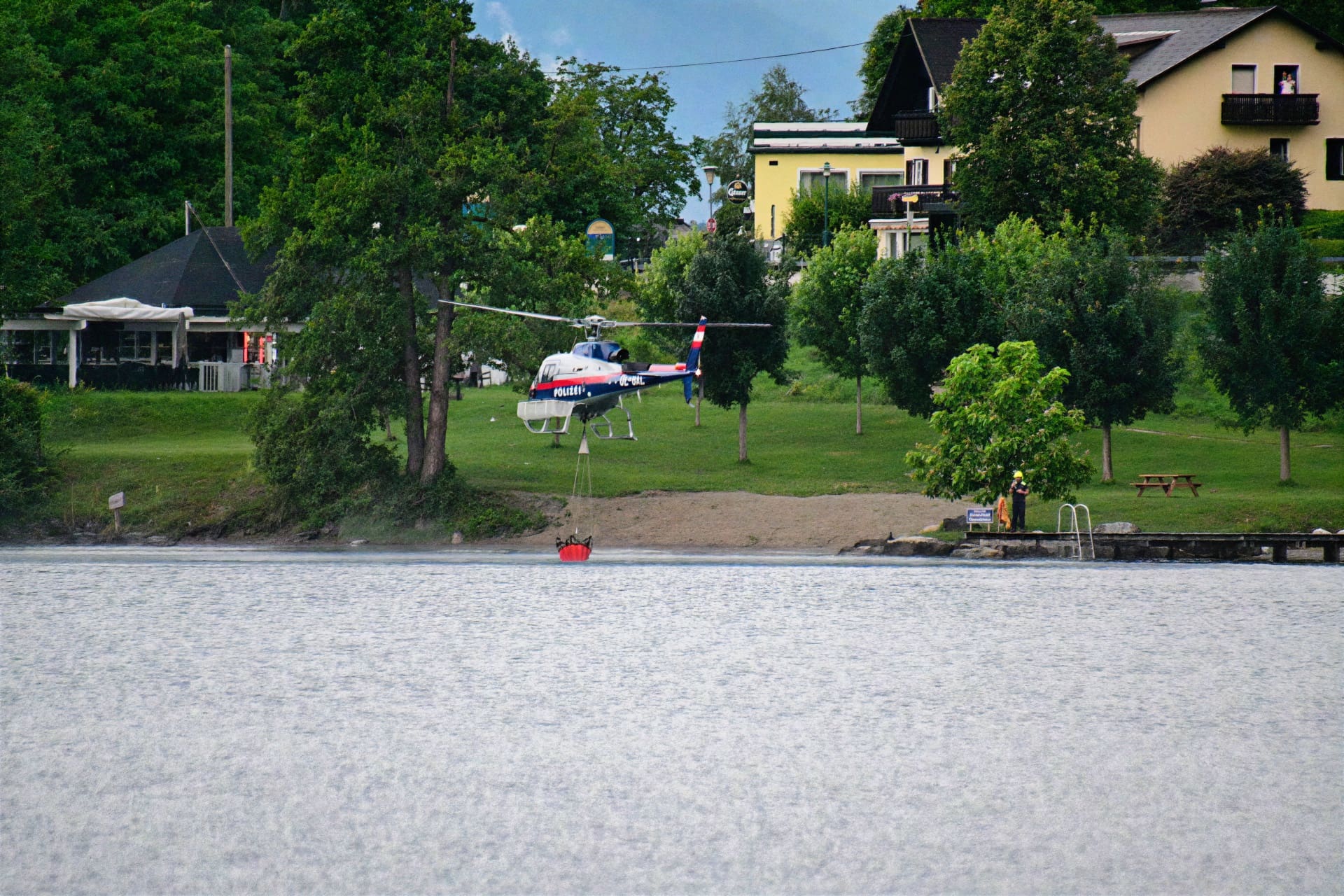 Polizeihubschrauber mit rotem Löschbehälter fliegt über See bei Pesenthein