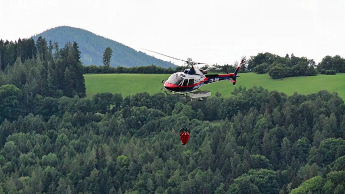 Polizeihubschrauber mit rotem Löschbehälter fliegt über dichtem Waldgebiet