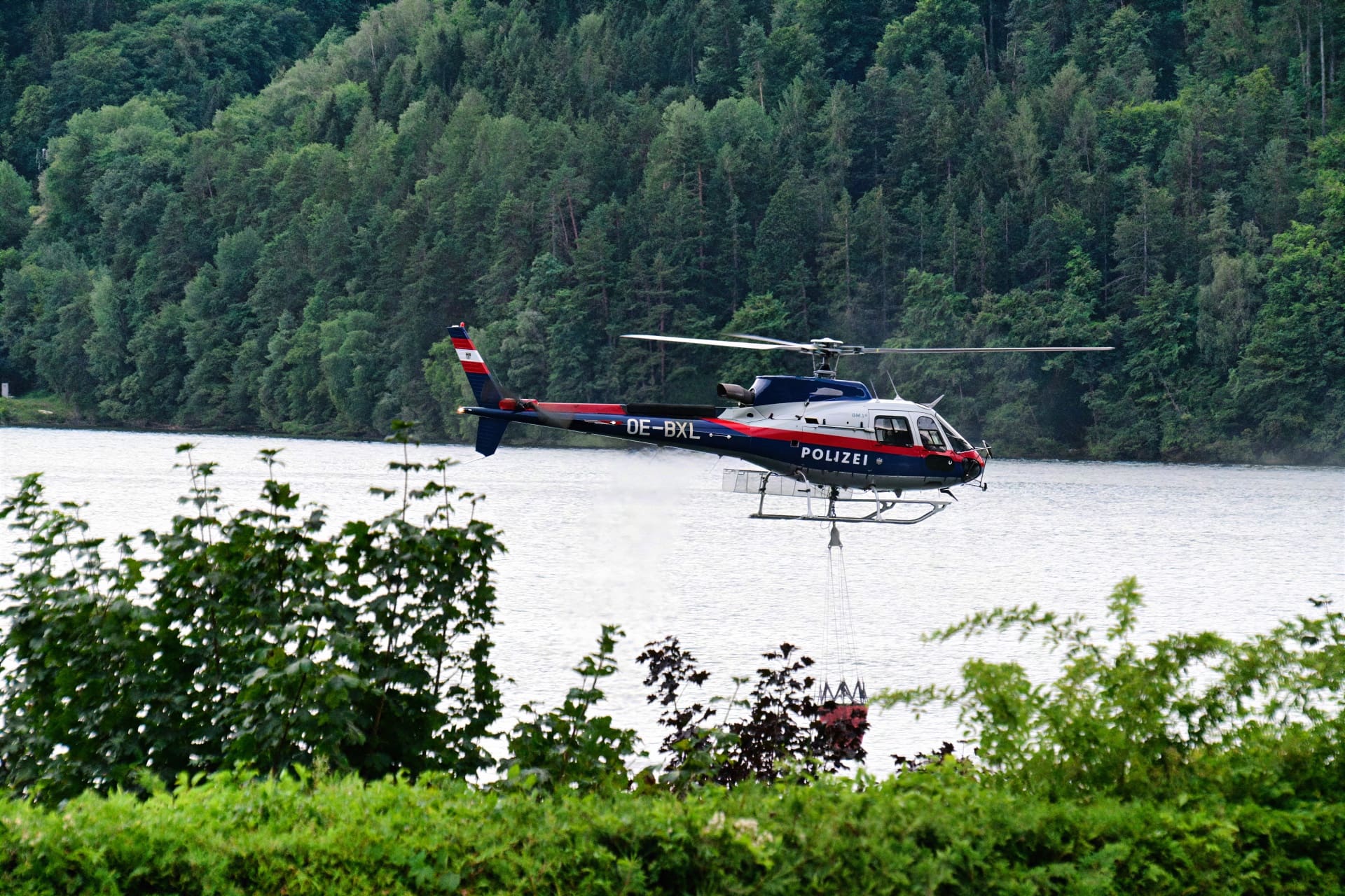 Polizeihubschrauber OE-BXL fliegt über dem Millstätter See vor bewaldetem Hang
