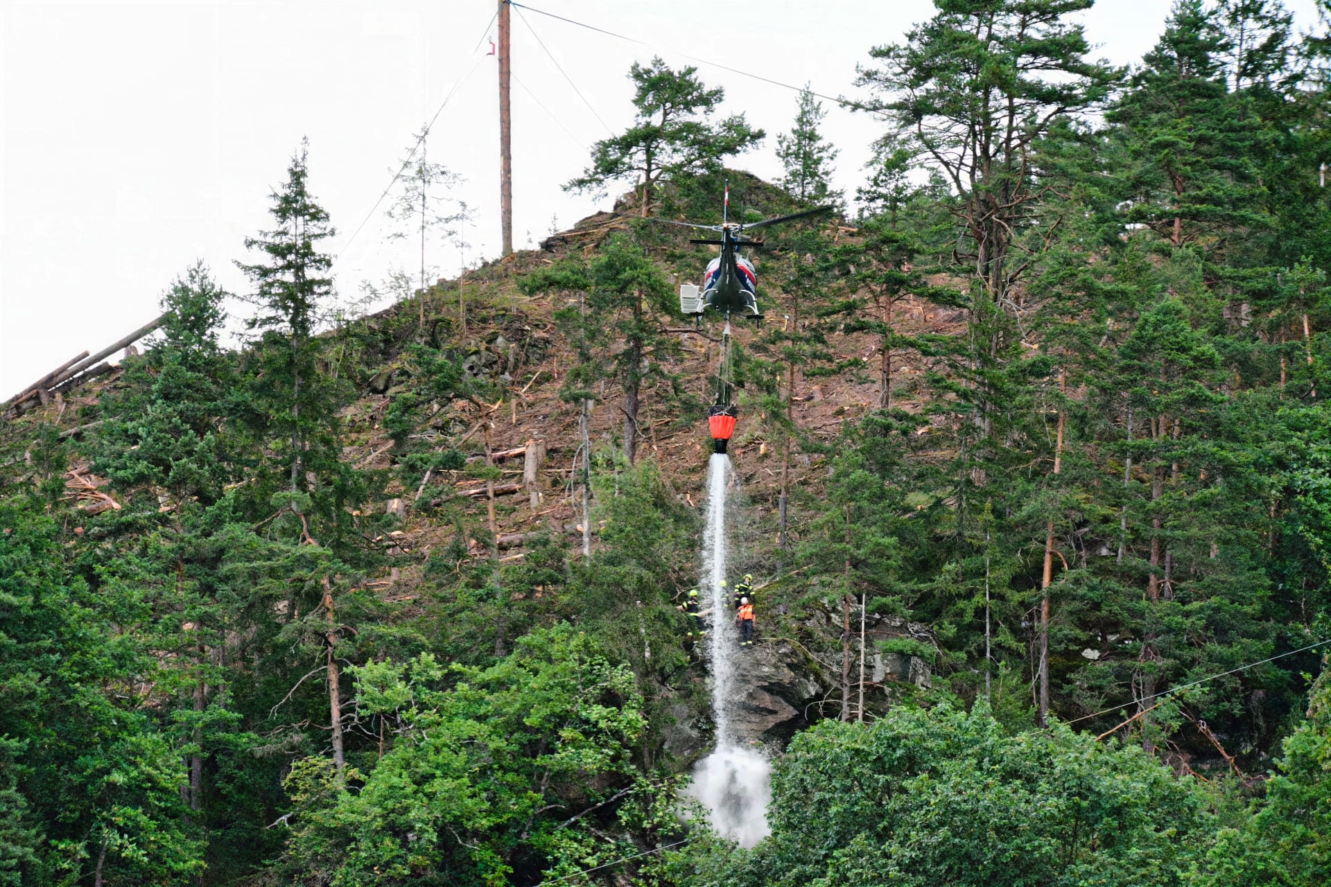 Hubschrauber mit rotem Löschbehälter fliegt über bewaldetem Hang mit Feuerwehrleuten