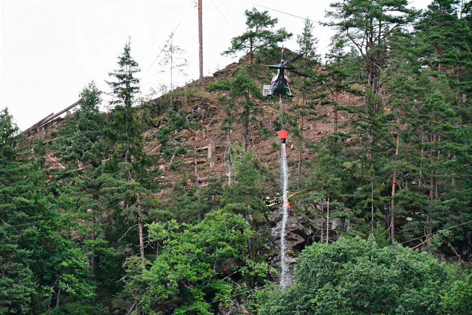 Hubschrauber mit rotem Löschwasserbehälter über Feuerwehrleuten auf bewaldetem Hang