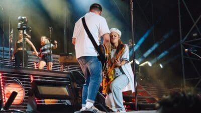 Ein Mann in weißem T-Shirt steht auf der Bühne neben einer sitzenden Gitarristin mit orangefarbener E-Gitarre.